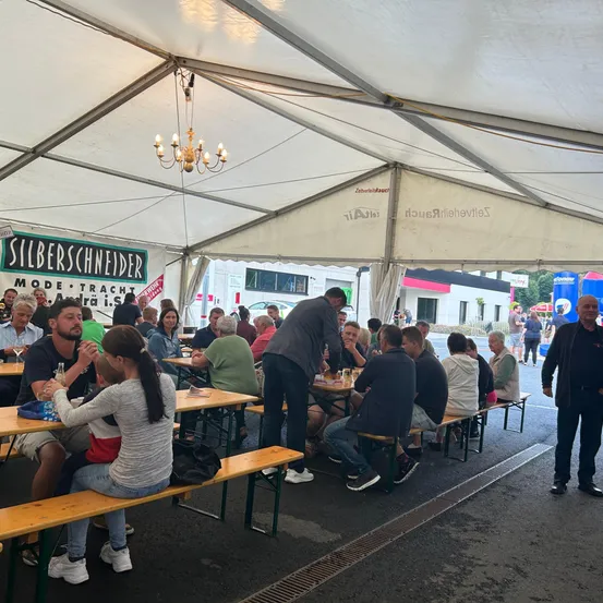 Ein Outdoor-Essbereich unter einem großen Zelt, mit Menschen, die an Tischen sitzen, und einem Mann, der vor einem steht. Eine Laterne hängt von der Decke.
