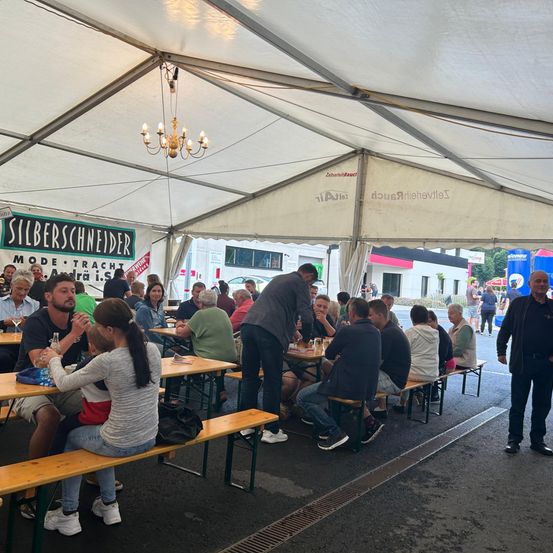 Ein Outdoor-Essbereich unter einem großen Zelt, mit Menschen, die an Tischen sitzen, und einem Mann, der vor einem steht. Eine Laterne hängt von der Decke.