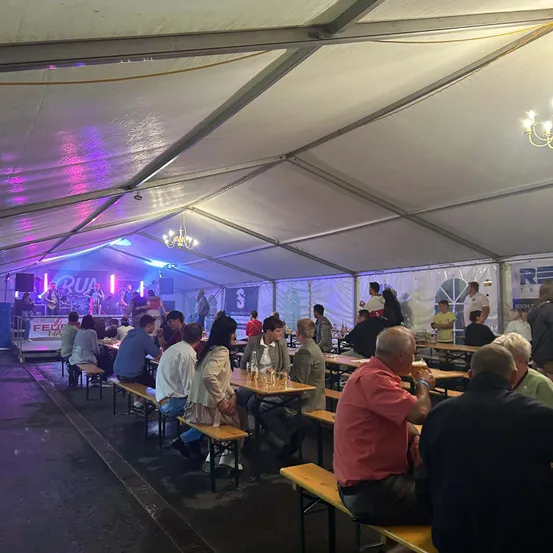 Ein Zelt mit Menschen an Tischen und Stühlen, beleuchtet von lila Lichtern. Eine Band spielt auf der Bühne vorne.
