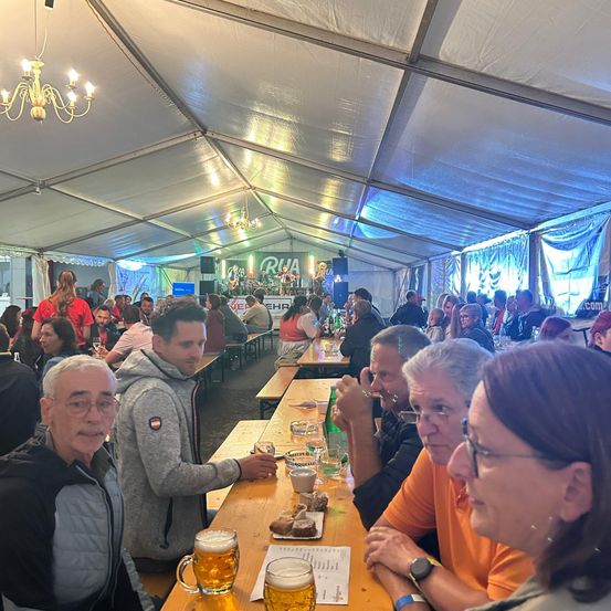 In einem Zeltrestaurant sitzen eine Gruppe von Menschen an Tischen und essen und trinken. Einige tragen Brillen und Jacken. Es gibt Gläser mit Bier und Essen auf den Tischen. Das Zelt hat eine Kronleuchter und Beleuchtung.