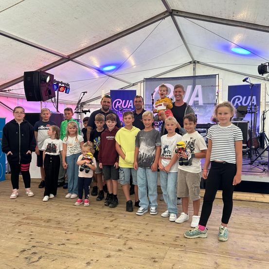 Eine Gruppe von Kindern und zwei Erwachsenen steht auf einer Bühne unter einem Zelt. Ein Erwachsener hält ein Baby. Musikinstrumente stehen hinter ihnen auf der Bühne.