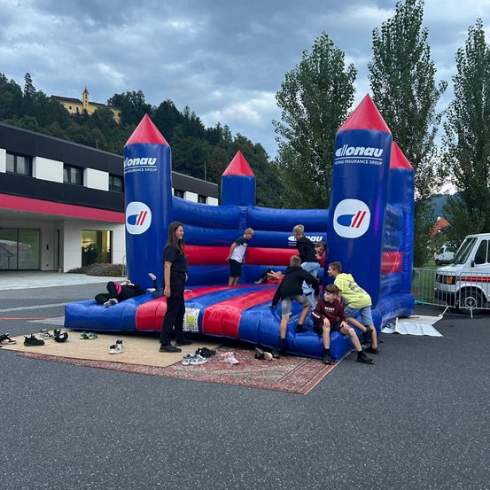 Eine Gruppe von Kindern und einer erwachsenen Frau spielen auf einem großen blauen und roten Sprungburg mit dem Wort Allonau darauf. Die Burg steht auf einem Teppich vor einem Gebäude.