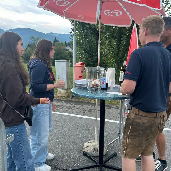 Zwei Frauen und zwei Männer stehen um einen kleinen Tisch mit einem roten Regenschirm, Flaschen und einem Mülleimer herum. Die Frauen tragen Jeans und Jacken, während die Männer Shorts und blaue Hemden tragen. Im Hintergrund befinden sich Bäume, ein Haus und Berge.