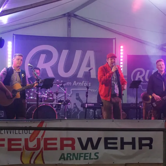 Eine Band tritt auf einer Bühne mit lila Beleuchtung auf, bestehend aus einem Gitarristen, einem Schlagzeuger, einem Keyboarder und einem Sänger in einer roten Jacke. Ein Banner zeigt 'RUA' und ein weiteres 'Feuerwehr Arnfels'.