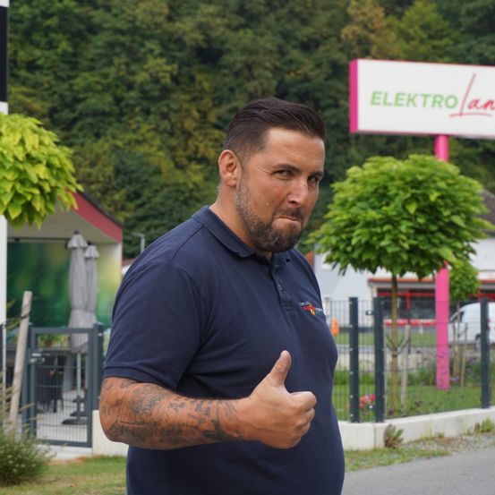 Ein Mann mit Tattoos am Arm gibt ein Daumen hoch vor einem Geschäft mit einem rosa Schild, auf dem Elektro Lan steht. Dahinter befinden sich Bäume, ein Zaun und ein Gebäude mit einem Schirm.