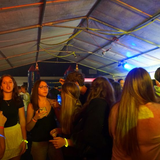 Eine Gruppe von Menschen ist drinnen versammelt, wahrscheinlich bei einem Festival oder einer Party. Einige halten Handys und andere schauen sich um. Der Raum ist mit bunten Lichtern beleuchtet.