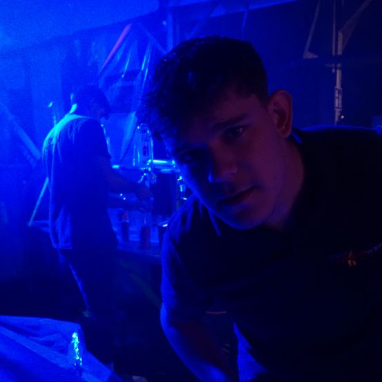 Ein Mann in einem schwarzen T-Shirt steht in einem schwach beleuchteten Raum mit blauen Lichtern. Hinter ihm steht ein weiterer Mann, der sich möglicherweise auf eine Aufführung vorbereitet.