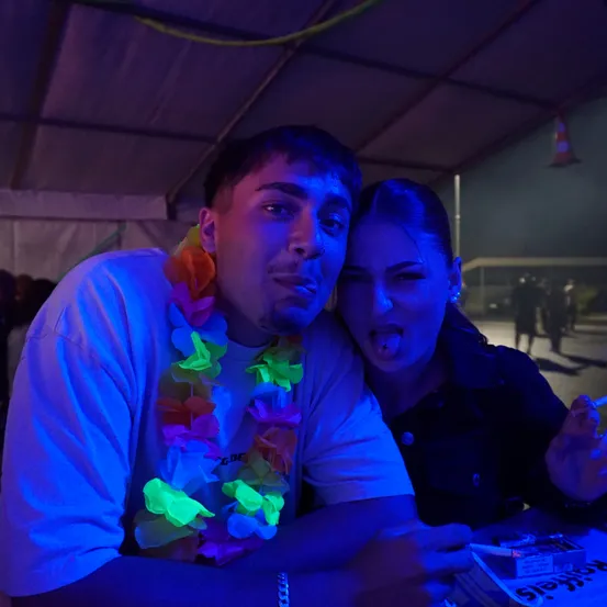 Ein junger Mann mit einer bunten Kette und einer Frau, die ihre Zunge herausstreckt, sitzen unter einem Zelt, möglicherweise auf einer Party, mit einem verschwommenen Hintergrund.