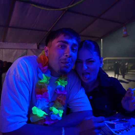 Ein junger Mann mit einer bunten Kette und einer Frau, die ihre Zunge herausstreckt, sitzen unter einem Zelt, möglicherweise auf einer Party, mit einem verschwommenen Hintergrund.