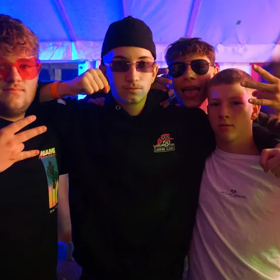 Vier junge Männer posieren für ein Foto unter Neonlichtern. Der Mann in der Mitte trägt eine schwarze Kapuzenjacke mit Logo. Die anderen tragen Sonnenbrillen und machen Gesten.