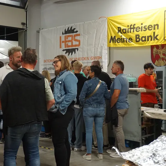 Eine Gruppe von Menschen ist in einem Raum versammelt. Einige tragen Brillen, andere lächeln. Ein Banner mit dem Text Raiffeisen Meine Bank hängt an der Wand. Ein Mann steht hinter einem Tisch mit einer Tasse darauf.