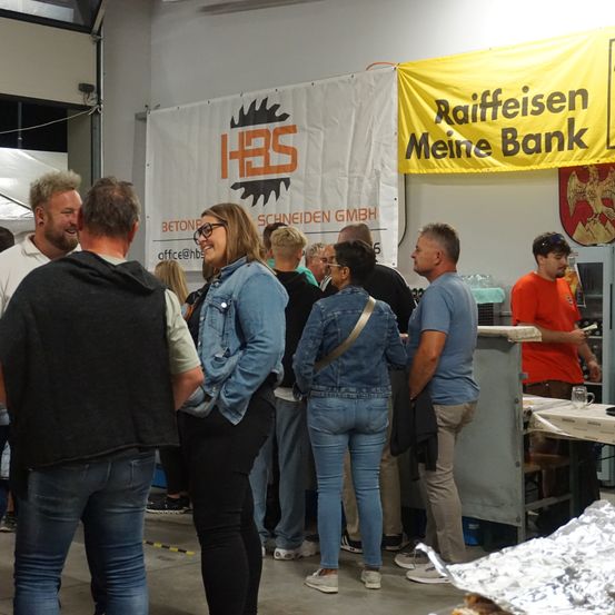 Eine Gruppe von Menschen ist in einem Raum versammelt. Einige tragen Brillen, andere lächeln. Ein Banner mit dem Text Raiffeisen Meine Bank hängt an der Wand. Ein Mann steht hinter einem Tisch mit einer Tasse darauf.