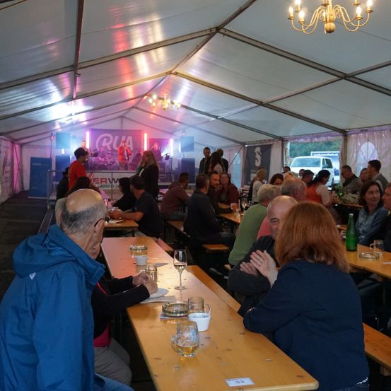 Eine Gruppe von Menschen sitzt in einem Zelt mit Bühne und Banner, wahrscheinlich bei einer Veranstaltung. Die Tische sind mit Weingläsern, Tassen und Tellern gedeckt. Ein Mann mit Brille schreibt, während eine Frau mit roten Haaren lächelt.