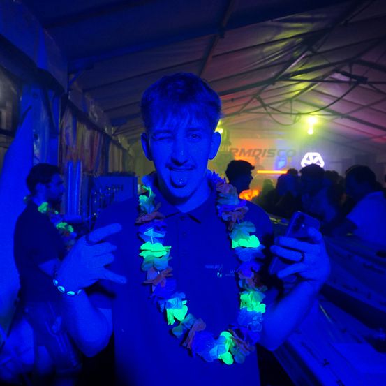 Ein Mann in einem blauen Shirt und einer Girlande mit leuchtenden Blumen posiert mit einem Handy bei einer leuchtenden Veranstaltung.