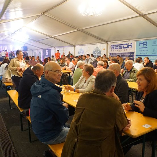 Viele Menschen sitzen in einem Zelt mit weißen Wänden und Dach. Einige unterhalten sich und essen, während andere stehen. An den Wänden hängen Banner.