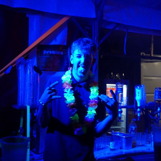 Ein Mann mit einem Blumenkranz steht an einer Bartheke unter blauer Beleuchtung. Verschiedene Flaschen und Gläser sind auf der Theke angeordnet.