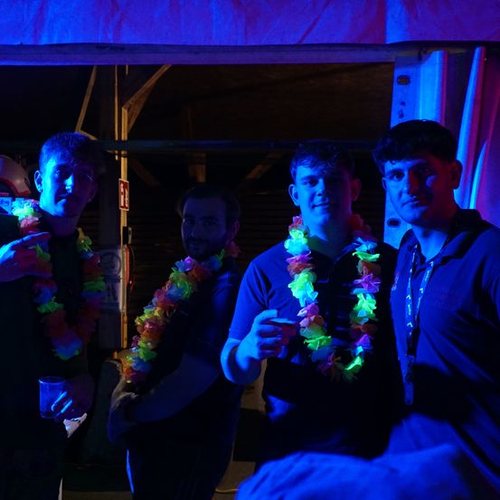 Vier Männer tragen farbenfrohe Blumenketten, die von blauen Lichtern beleuchtet werden. Sie stehen in einem dunklen Bereich, wahrscheinlich auf einer Party.