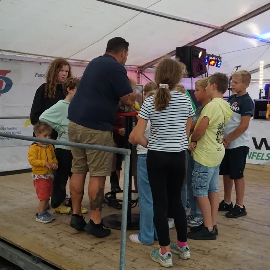 Eine Gruppe von Kindern und Erwachsenen steht um einen Tisch herum, wahrscheinlich bei einer Veranstaltung. Der Mann schaut auf den Tisch, während andere zusehen. Die Bühne ist aus Holz mit einem weißen Zelt darüber.