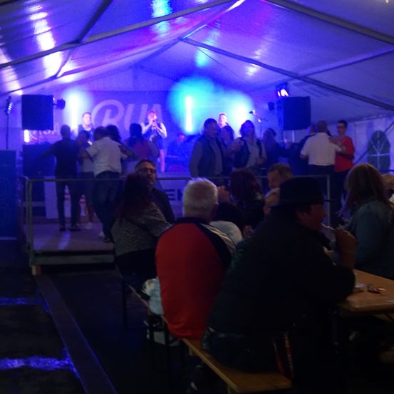 Eine Gruppe von Menschen tanzt in einem Zelt mit lila und blauer Beleuchtung. Einige Personen stehen auf der Bühne, andere sitzen an Tischen.