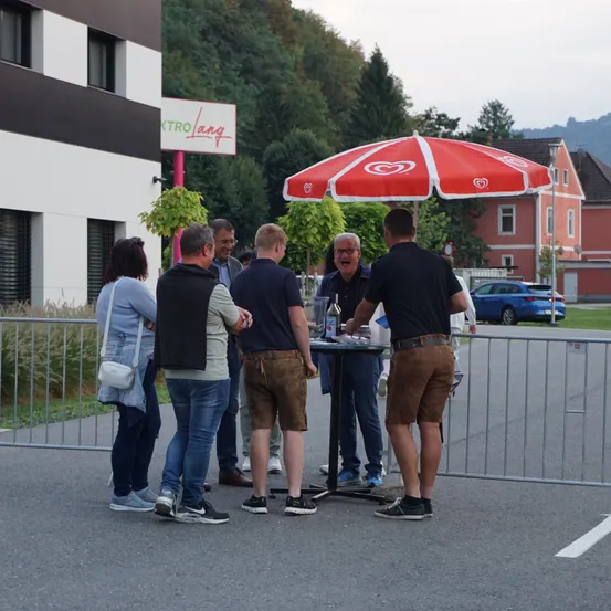 Eine Gruppe von Menschen steht um einen Tisch unter einem roten Schirm. Sie befinden sich vor einem Gebäude mit dem Schild KTRo Lang. Im Hintergrund gibt es Bäume und ein Haus. Ein blaues Auto ist in der Nähe geparkt.