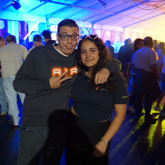 Ein Mann und eine Frau posieren für ein Foto bei einer lebhaften Veranstaltung, umgeben von anderen Gästen unter farbiger Beleuchtung.