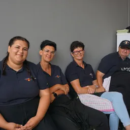 Vier Erwachsene in schwarzen Shirts und Schürzen sitzen in einer Reihe und lächeln für die Kamera. Die Frau rechts trägt eine Brille und einen Hut.
