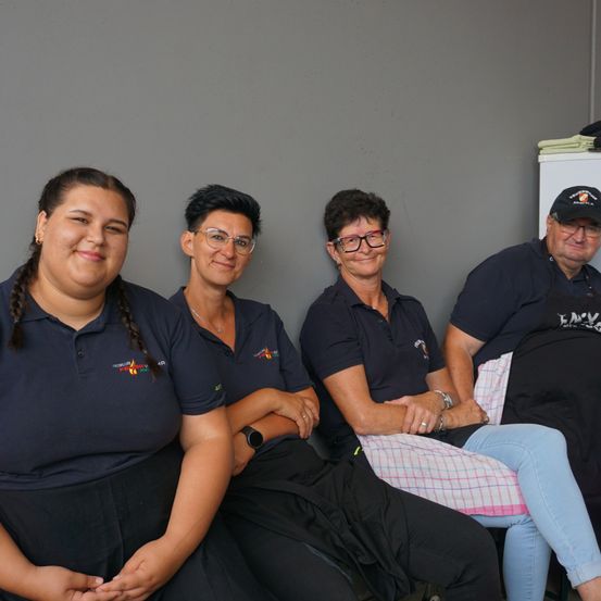 Vier Erwachsene in schwarzen Shirts und Schürzen sitzen in einer Reihe und lächeln für die Kamera. Die Frau rechts trägt eine Brille und einen Hut.
