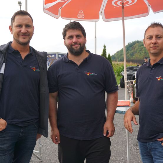 Drei Männer stehen draußen, zwei in blauen Poloshirts mit Logos und einer mit einer Jacke. Sie lächeln und stehen unter einem roten Regenschirm. Hinter ihnen steht ein Tisch mit einem Glas und einer Flasche, und in der Ferne ist ein Gebäude zu sehen.