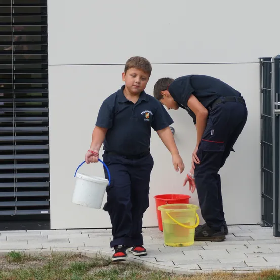 Ein junger Junge in einer Marineuniform hält einen weißen Eimer und ein anderer Junge wäscht seine Hände mit Wasser aus einem Wasserhahn.