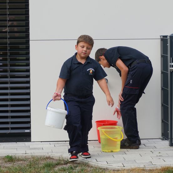 Ein junger Junge in einer Marineuniform hält einen weißen Eimer und ein anderer Junge wäscht seine Hände mit Wasser aus einem Wasserhahn.