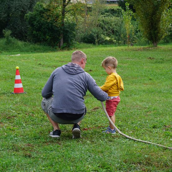 Ein Mann kniet auf dem Gras und hält die Hand eines Kindes, während das Kind einen Schlauch hält. Sie befinden sich in einem Park mit Bäumen und einem Verkehrskegel.
