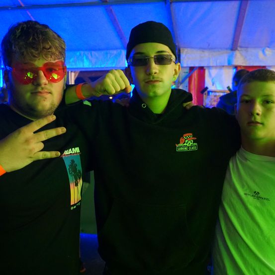 Drei junge Männer stehen unter einem Zelt mit blauer Beleuchtung. Einer trägt rote Sonnenbrille, ein anderer einen schwarzen Hut und der dritte ein grünes Shirt.