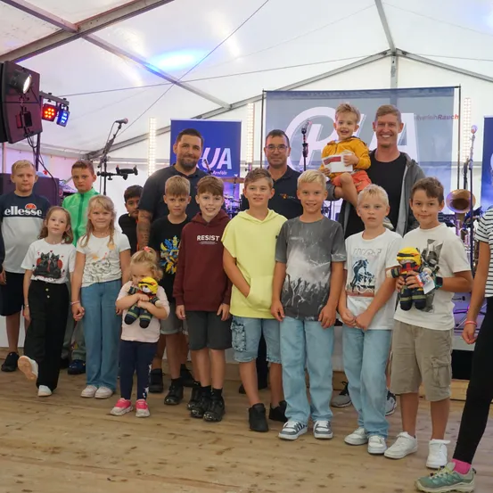 Eine Gruppe von Kindern und Erwachsenen, möglicherweise Musiker, posiert für ein Foto in einem Zelt. Der Mann in der Mitte hält ein Baby. Einige von ihnen halten Plüschtiere.