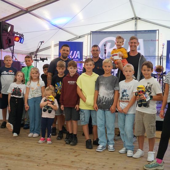 Eine Gruppe von Kindern und Erwachsenen, möglicherweise Musiker, posiert für ein Foto in einem Zelt. Der Mann in der Mitte hält ein Baby. Einige von ihnen halten Plüschtiere.