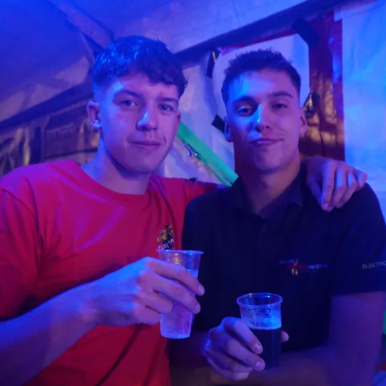 Zwei junge Männer posieren für ein Foto in einem schwach beleuchteten Raum und halten beide Getränke in Gläsern. Der Mann auf der linken Seite trägt ein rotes T-Shirt, während der Mann auf der rechten Seite ein schwarzes Polohemd mit Logo trägt.