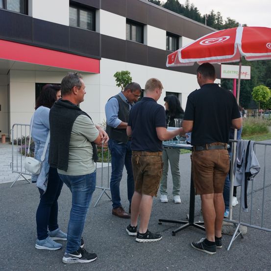 Eine Gruppe von Menschen steht vor einem Gebäude in der Nähe eines Tisches mit einem Schirm. Sie scheinen auf etwas zu warten.