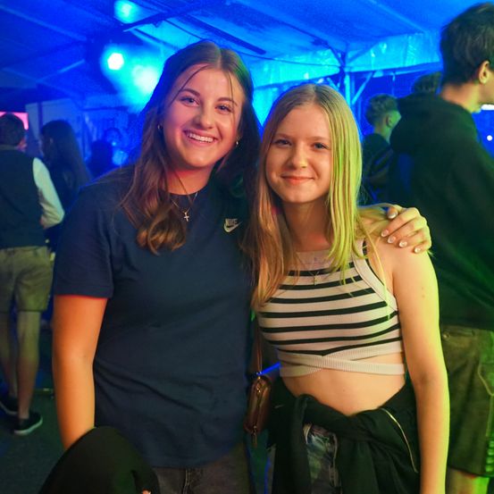 Zwei junge Frauen lächeln für die Kamera bei einer Clubveranstaltung. Eine trägt ein schwarzes Nike-Shirt, die andere ein gestreiftes Crop-Top. Verschwommene Figuren im Hintergrund.
