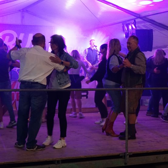 Unter einem lila Licht tanzen eine Gruppe von Menschen auf der Bühne, mit einem Mann, der im Hintergrund die Gitarre spielt.