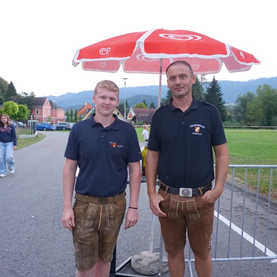 Zwei Männer stehen vor einem roten Regenschirm. Einer trägt braune Shorts und der andere ein schwarzes Shirt. Dahinter befindet sich eine Wiese mit Bäumen und Häusern.