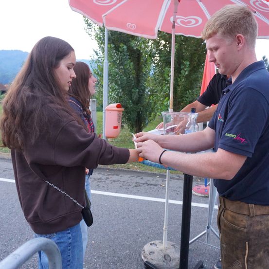 Eine Frau reicht einem Mann einen Gegenstand an einem Tisch unter einem rosa Regenschirm. Dahinter hält eine Person eine rote Flagge. Im Hintergrund befindet sich eine Wiese und Berge.