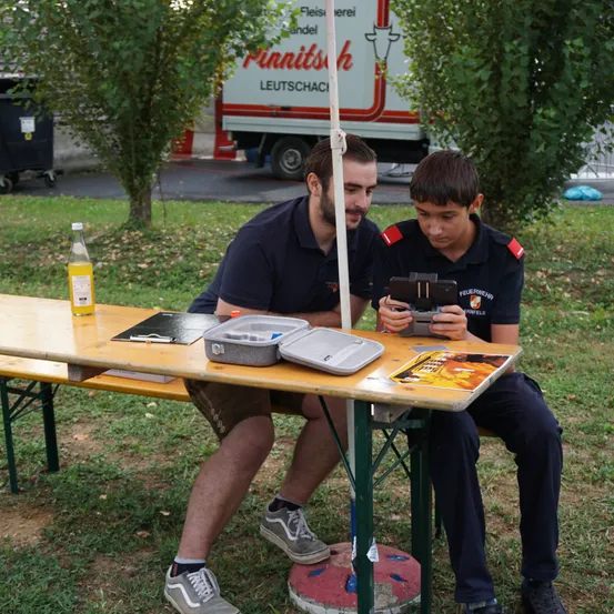 Zwei Männer sitzen auf einer Bank unter einem Baum. Ein Mann hält eine Kamera und der andere schaut auf ein Tablet. Eine Flasche und einige Papiere liegen auf dem Tisch.