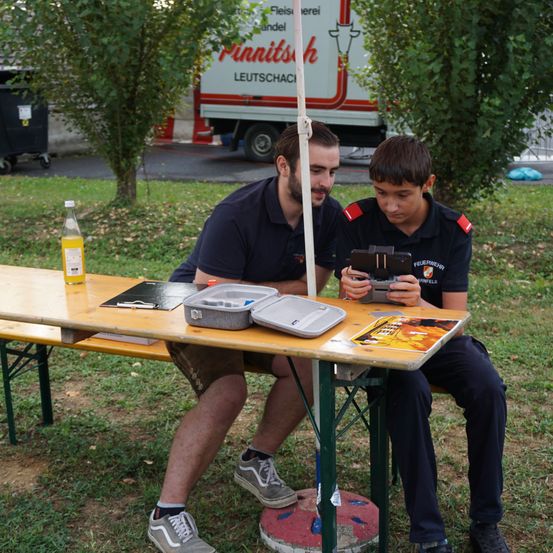 Zwei Männer sitzen auf einer Bank unter einem Baum. Ein Mann hält eine Kamera und der andere schaut auf ein Tablet. Eine Flasche und einige Papiere liegen auf dem Tisch.