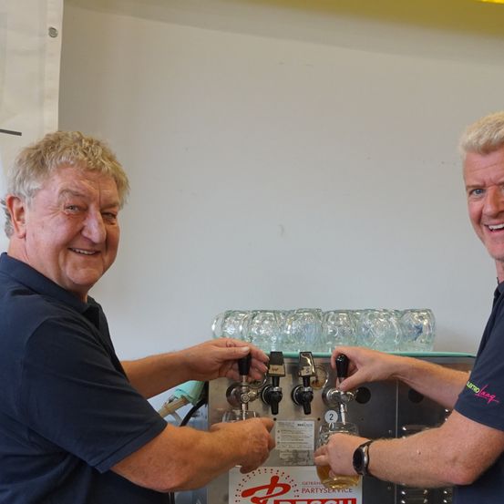 Zwei Männer lächeln und stehen hinter einer Bierzapfanlage. Sie tragen beide schwarze Polo-Shirts. Einer von ihnen hält ein Glas Bier in der Hand.