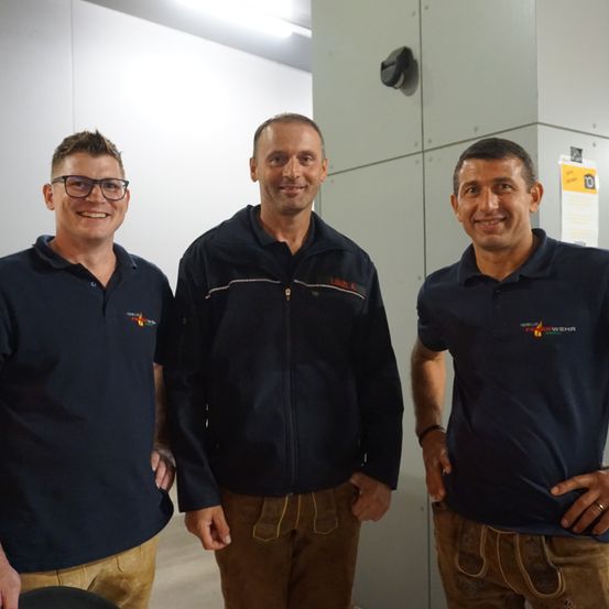 Drei Männer stehen zusammen in einem Raum mit weißer Wand und grauen Paneelen. Sie tragen blaue Hemden mit Logos, einer hat eine Brille und die anderen tragen braune Hosen.