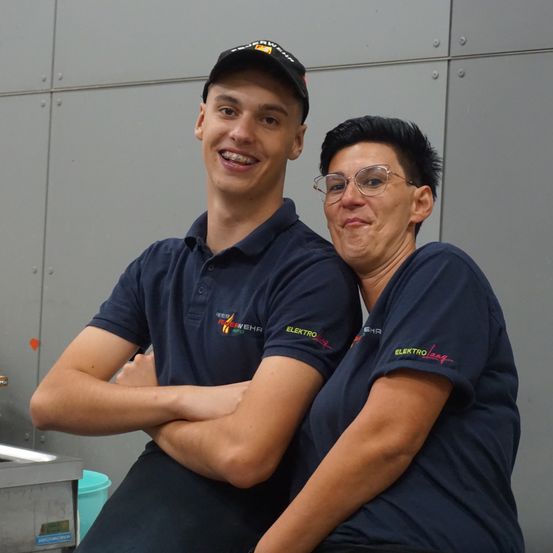 Zwei Personen in passenden dunkelblauen Hemden und einer Kappe lächeln und posieren für ein Foto. Sie scheinen in einer industriellen Umgebung zu stehen.