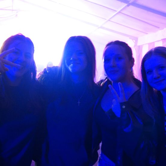 Vier Frauen stehen unter lila Licht. Die Frau auf der linken Seite macht ein Friedenszeichen, während die Frau in der Mitte eine Kreuzkette trägt. Die Frau auf der rechten Seite lächelt.