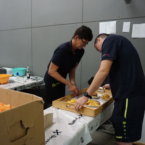 Zwei Personen bereiten Essen in einem Raum zu. Eine Frau mit Brille und Armband steht und ein Mann beugt sich über einen Tablett mit Essen. Ein Tisch hat Teller mit Essen, einen Topf und eine Flasche. Ein anderer Tisch hat Papiere, die an der Wand befestigt sind.