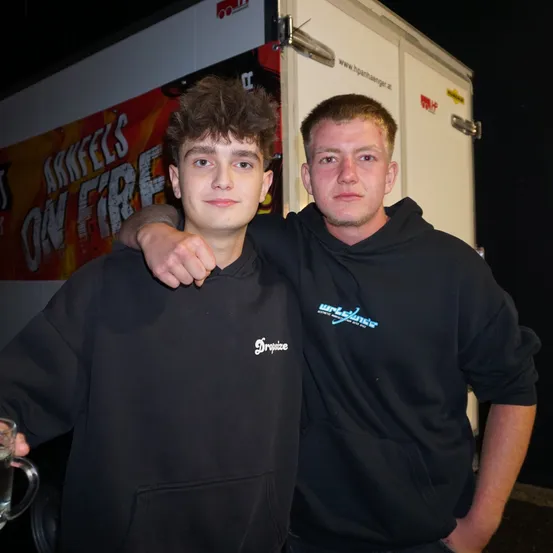 Zwei junge Männer stehen eng beieinander und lächeln für ein Foto. Einer hält ein Glas. Sie tragen schwarze Hoodies, wobei einer ein weißes Logo zeigt. Hinter ihnen steht ein weißer Anhänger mit einem roten Banner, auf dem 'ARKFEELS ON FIRE' steht.
