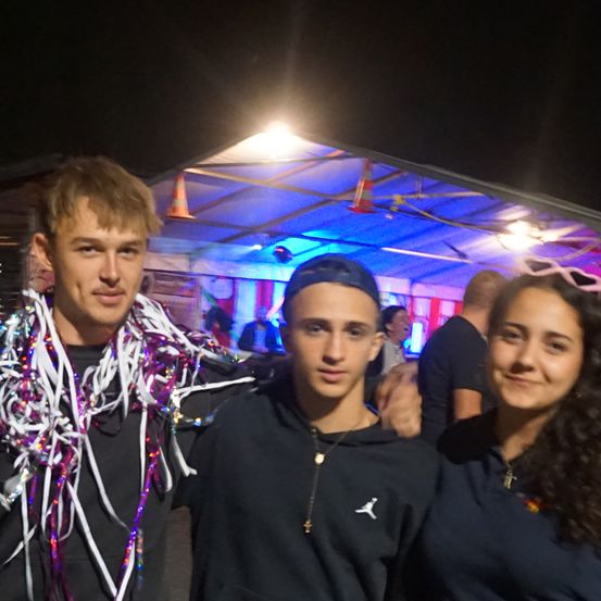 Drei Menschen stehen eng beieinander, zwei Männer und eine Frau, unter einem Zelt mit blauen Lichtern. Ein Mann trägt eine bunte Girlande, und ein anderer trägt eine Kappe. Eine Menschenmenge ist im Hintergrund schwach zu sehen.