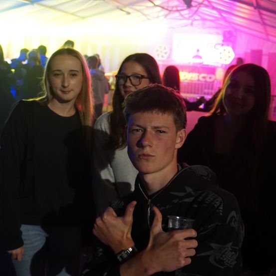 Eine Gruppe von Freunden steht bei einem Disco-Event zusammen. Ein junger Mann hält einen Becher in der Hand und macht eine Daumen-Hoch-Geste. Der Hintergrund zeigt einen schwach beleuchteten Raum mit einer Mischung aus gelben und lila Lichtern.